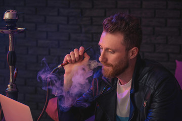 Young man smoking hookah while working on laptop in cafe