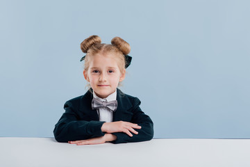 Wall Mural - education. little schoolgirl keep your hands on the table, look at the camera,  sitting at the white table, isolated on blue background, copy space, school concept