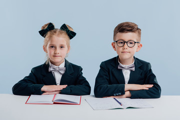 Wall Mural - education. two schoolgirls look at the camera, notebooks on the table, school uniform, boy and girl, sitting at the white table, isolated on blue background, copy space, school concept