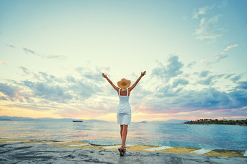 Traveling by Greece. Young happy woman rising hands up enjoying beautiful sunset on the sea.