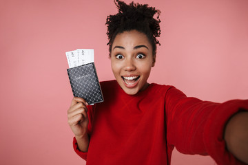 Wall Mural - Excited young african woman posing isolated over pink wall background take selfie by camera with passport and tickets.