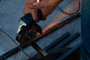 Close-up view strong man master without gloves on arms, performs metal cutting with an angle grinder in the garage workshop, blue and orange sparks fly to the sides