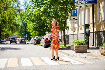 Wall Mural - Young beautiful blonde woman in a red flower dress crosses the road at a crosswalk