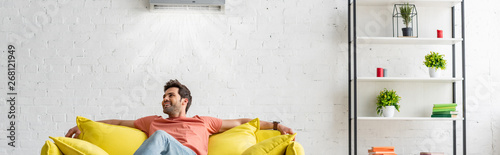 Fotografía panoramic shot of handsome man sitting on yellow sofa under air conditioner at h
