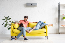 Happy Man With Smiling Girlfriend Relaxing On Yellow Sofa Under Air Conditioner At Home