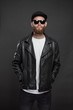 © 4Max - Man wearing leather biker jacket or asymmetric zip jacket with black cap, jeans and sunglasses. Handsome hipster man over gray background