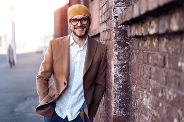 Portrait of stylish handsome young man in glasses with bristle standing outdoors. Man wearing jacket and shirt, leaning against wall.