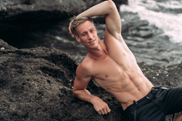 Wall Mural - Portrait of young man with naked torso standing on rock beach with black sand