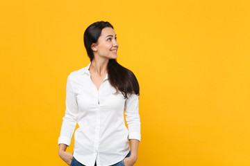 Wall Mural - Portrait of smiling young woman in white shirt standing, holding hands in pockets, looking aside isolated on yellow orange wall background in studio. People lifestyle concept. Mock up copy space.