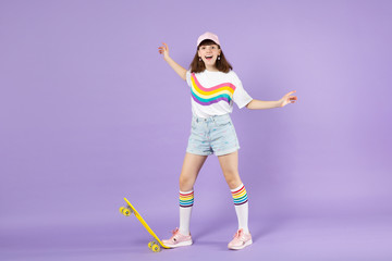 Wall Mural - Cheerful teen girl in vivid clothes standing with yellow skateboard spreading hands isolated on violet pastel wall background in studio. People sincere emotions, lifestyle concept. Mock up copy space.