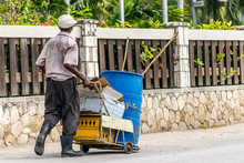Black Elderly Man Wearing Rubber Boots And Dirty Clothing On The Street Pushes Cart With Plastic Drum, Crate, Boxes And Sheets Of Cardboard Following A Clean Up Job. Labor/Labour Day Project.