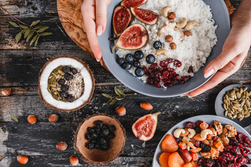 Wall Mural - Woman holding plate of Rice coconut porridge with figs, berries, nuts and coconut milk on rustic wooden background. Healthy breakfast ingredients. Clean eating, vegan food concept. Top view.