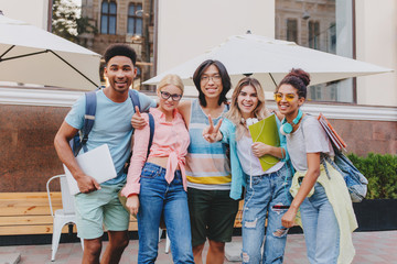 Wall Mural - Laughing asian boy in glasses and shorts embracing charming blonde girls in front of outdoor cafe. Joyful students came to open-air restaurant to celebrate end of exams