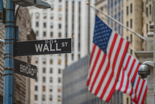 Wall Street "WALL ST" Sign And Broadway Street Over American National Flags In Front Of NYSE Stock Market Exchange Building Background. The New York Stock Exchange Locate In Economy District