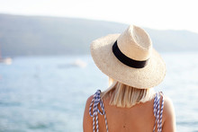 Woman In Straw Hat At Sea Beach In Summer Vacation. Female Tourist Is Enjoying Holiday, Travel And Summertime. Girl Is Looking At View Of Ocean, Island, Mountains.