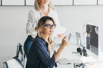 Pretty freelancer girl sitting near computer with black screen and looking away with smile. Indoor portrait of european blonde girl in white shirt holding documents beside colleague's table.