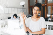 Glad african woman in white t-shirt posing with okay sign in front of busy colleague. Indoor portrait of excited black girl having fun in office during break.