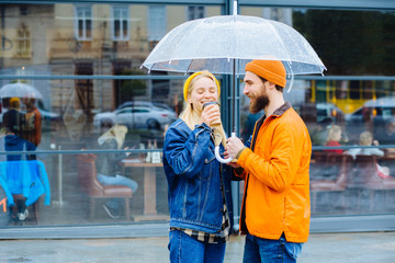 Happy hipster couple hugging drinking warm coffee under trensparent umbrella in rainy spring cold weather urban city street on background.