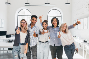 Wall Mural - Blonde female student in ripped jeans holding laptop and laughing after hard test. Asian guy in glasses waving hands, celebrating end of exams and beginning of vacation.