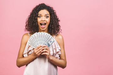 Rich girl! Money winner! Surprised beautiful african american woman in dress holding money and looking at the camera isolated against pink background.