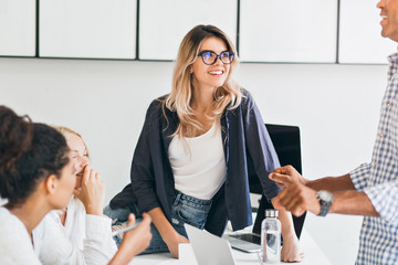 Wall Mural - European female student in glasses chilling on table and listening university friend in wristwatch. African young woman talking with collegues in office during coffee break.