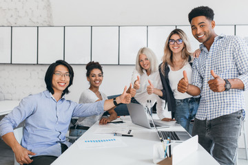 Wall Mural - Blonde secretary sitting on table while office workers posing with thumbs up. Indoor portrait of happy asian manager in trendy shirt smiling in conference hall with foreign partners.