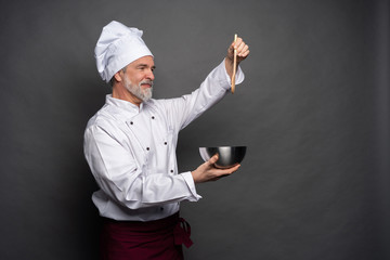 Wall Mural - Smiling mature male chef with bowl and cooking vane in hands on black background.