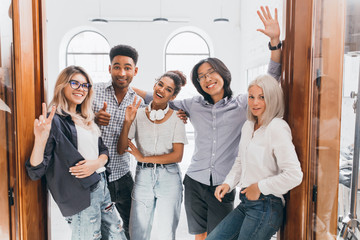 Wall Mural - African young man with surprised smile standing beside doors with colleagues after work day. Asian student in glasses posing with university friends near library.