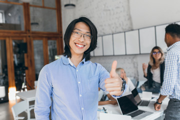 Asian laughing boy posing with thumb up at the beginning of the workday. Chinese office worker in blue shirt and glasses smiling with laptop on background.