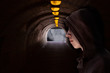 © Ole - Boy with hood standing in a dark underground passage with yellow light bulbs at the roof and light at the end. Dangerous youth concept.