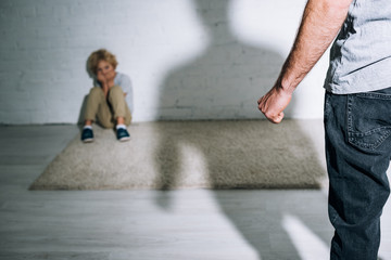 partial view of abusive father and scared son sitting on carpet