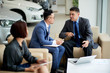 © DragonImages - Businessmen sitting on sofa and discussing a purchase of new car with businesswoman sitting near by them and listening to their conversation  in car showroom