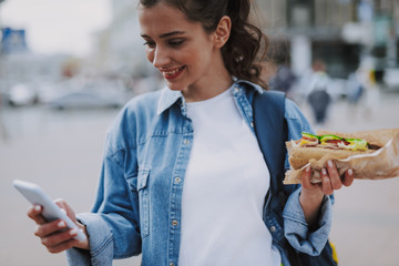 Pretty female checking mobile with hot dog in hand