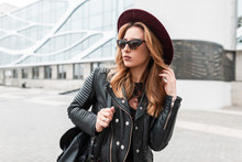Sexy Modern Hipster Woman In A Luxurious Purple Hat In A Fashionable Black Jacket With A Backpack In Stylish Sunglasses Walks Along A Street In The City Near A Vintage Building. Cute Girl Outdoors.