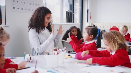 Wall Mural - Young female infant school teacher sitting at a table in a class with her pupils, counting with girl