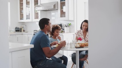 Wall Mural - Young family sitting at table in the kitchen eating together, selective focus, panning shot