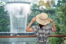 young woman wear blue dress and hat, Asian traveler standing and looking to beautiful rain vortex at Jewel Changi Airport, landmark and popular for tourist attractions in Singapore. Travel concept