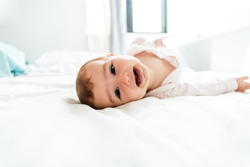 Wall Mural - Happy and smiling adorable 6 month old baby girl lying on a bed, lifestyle isolated on natural white background.