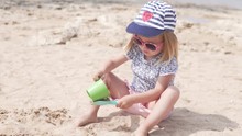 Kid Playing With Sand. Little Girl Plays On The Beach. Fashionable Little Girl Pours Sand From The Bucket To The Spatula