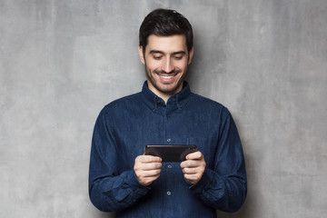 Poster - Handsome smiling guy using his smartphone for gaming or watching video isolated on gray textured wall background