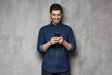 Wall Mural - Young man smiling and laughing as he is browsing Internet or communicating via phone, isolated on gray textured wall