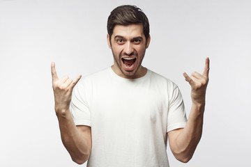 Wall Mural - Young man showing rock sign with his fingers as if listening to favourite music, isolated on white background