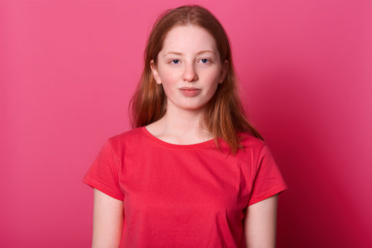 Half lengh portrait of young student female looks with calm facial expression at camera, wearing red casual t shirt, has long straight brown hair and beautiful blue eyes, isolated over pink background