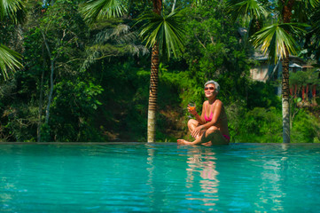 Wall Mural - natural lifestyle portrait of attractive and happy middle aged Asian woman relaxed at tropical resort infinity simming pool with jungle background enjoying a drink relaxed