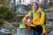 Glad tourist hold coffeepot, has picnic on stump, wears scarf on head, yellow raincoat, smiles positively, carries rucksack, poses againt blurred forest background on sunny day. Woman at tourist camp