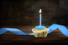Muffins Or Cupcakes With A Blue Burning Candle And A Ribbon On A Dark Wooden Board Against A Brown Rustic Background, Birthday Concept With Copy Space