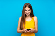 © luismolinero - Young woman with long hair over isolated blue wall sending a message with the mobile