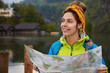 © wayhome.studio  - Positive young woman with joyful expression, holds location map, stands against beautiful nature with river, small houses and forest, enjoys leisure, dressed in anorak and headband, has smile on face