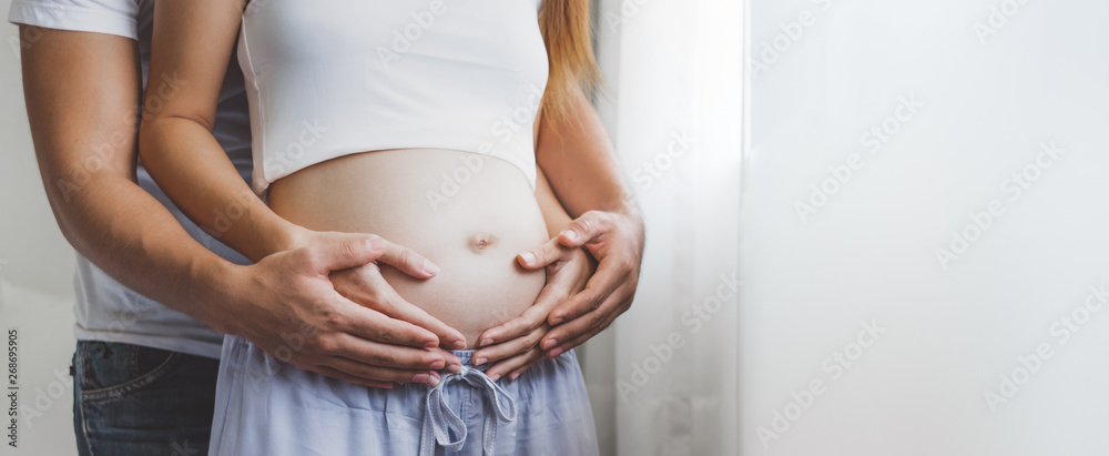 Young pregnant woman holding her belly with husband over white window with copy space banner - obrazy, fototapety, plakaty 