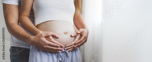 Fotografía Young pregnant woman holding her belly with husband over white window with copy